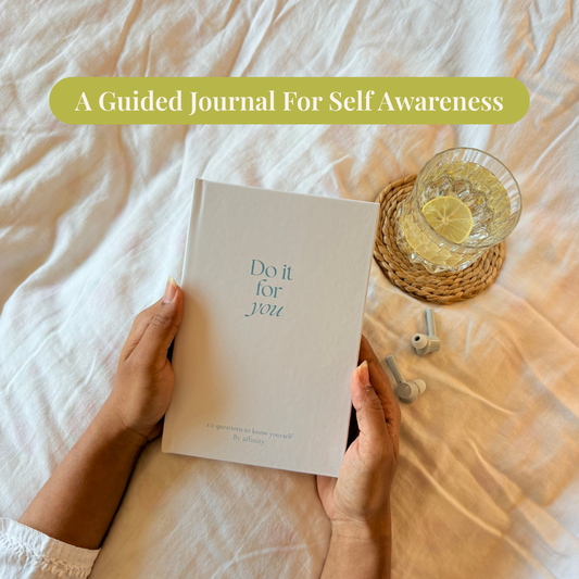 Person holding a guided journal self reflection journal for self awareness on a white bedsheet with grey airpods and water in a glass with a sliced lime on a coaster.