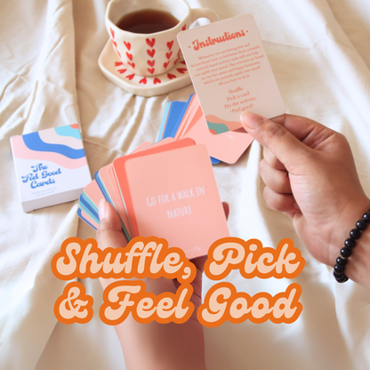 Person holding a card from a deck of feel good cards with a cup of tea and additional cards on a light surface.